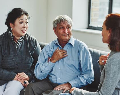 A lady sits with her dad who is smiling and placing a hand on his chest as if gesturing to himself.